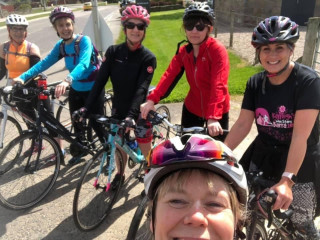 Group of female cyclists gathered together by the side of a road, smiling for the camera. In the foreground the photographer is captured in a 'selfie' pose
