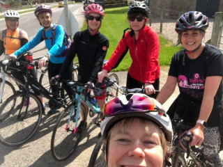 Group of female cyclists gathered together by the side of a road, smiling for the camera. In the foreground the photographer is captured in a 'selfie' pose