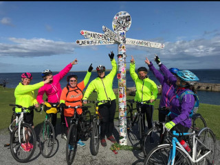 Group of women on bikes reaching for the sign at John O'Groats on a beautiful sunny day