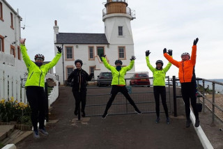 Group of female cyclists, jumping in the air, in the background is a lighthouse building