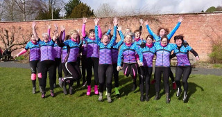 Group of female cyclists jumping in the air. All of the cyclists are wearing the same club top, in blue and pink with stripes and spots