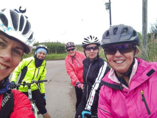 Jenny taking a selfie on a Breeze ride, to the right of her are four women smiling for the camera. All the women are wearing cycle helmets and rain jackets. The picture looks to have been taken at the side of a road in the countryside 