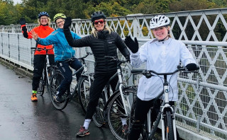 Group of female cyclists on a bridge, with their bikes, smiling for the camera. In the background are trees in full bloom