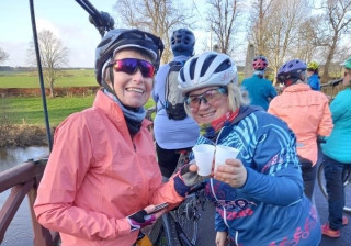Two female cyclists standing on a bridge, making a toast with plastic cups. In the background other female cyclists can be seen milling about