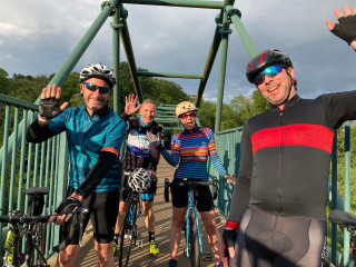 Image of four adult cyclists standing on a bridge with their bikes, waving at the camera
