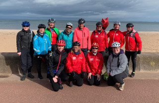 A group of 12 female cyclists posing for a photograph beside a beach. Five of the group are wearing Breeze Champion kit. One of the group is holding a red balloon in the shape of a star