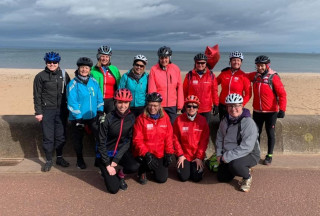 Group of female cyclists in front of a wall at Portobello Beach. Eight women are standing, four women are crouched in front of them. In the background there is sand, sea and sky.