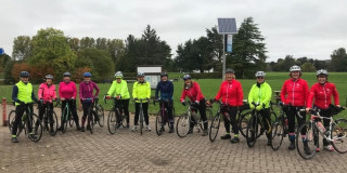 Group of female cyclists, standing in a row, with their bikes. In the background is trees and an overcast sky