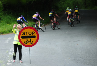 A female marshal holding a 'Stop! Cycle Race' sign as riders pass behind