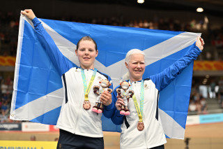 Alieen (R) with Ellie Stone (L) on the podium with the salitre held aloft after winning bronze at the 2022 Commonwealth Games. Both medals are round their necks and they are holding their toy plushy